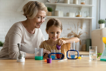 Obraz na płótnie Canvas Happy caring middle aged older grandmother doing chemical tests with joyful candid little preschool grandchild in modern kitchen. Smiling curious kid girl studying with pleasant granny in kitchen.