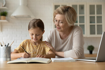 Adorable attentive small kid girl sitting at table, doing homework with caring older mature grandmother. Pleasant middle aged woman helping little child preparing for exams, homeschooling concept.