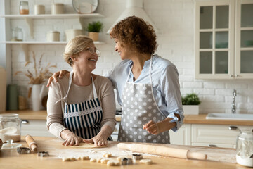 Smiling affectionate young woman cuddling loving older mature retired mother, enjoying cooking...
