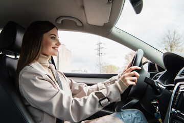side view of smiling woman in trench coat driving car in city