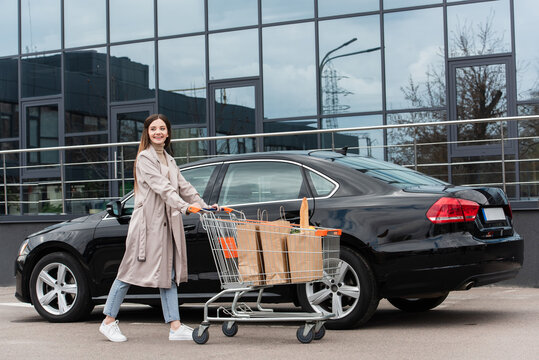 Cheerful Woman Walking With Shopping Trolley Near Black Car On Parking