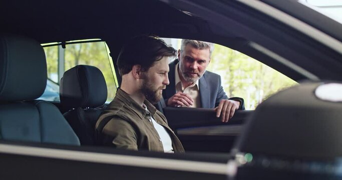Showroom car, caucasian buyer sit in automobile and test it. Bearded salesnmanager car agent in suit help the client.