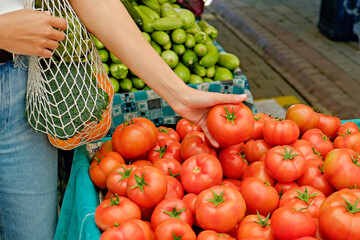 Plant based diet concept. Young woman with reusable eco friendly net bag picking fresh tomatoes on farmers market. Conscious shopping for organic local vegetables. Close up, copy space, background.