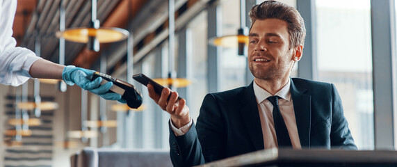 Handsome young smiling man in full suit making a contactless payment while sitting in the restaurant