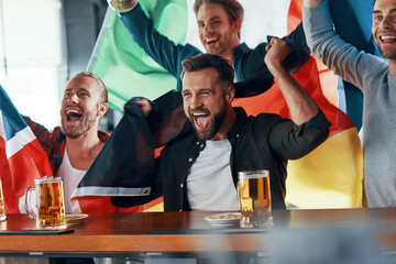 Cheering young men covered in international flags enjoying beer while watching sport game in the pub