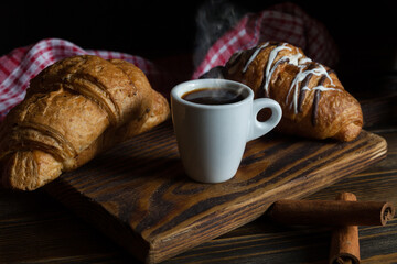 A mug of aromatic coffee in a beautiful mug with a croissant and cinnamon sticks. Dark background. 