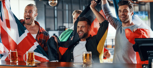 Cheering young men covered in international flags enjoying beer while watching sport game in the pub