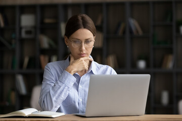 Fototapeta premium Thoughtful young businesswoman employee entrepreneur on eyeglasses looking at computer screen, pondering problem solution, creating startup project ideas or developing strategy in office room.