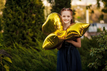 A beautiful little girl with bright yellow balloons in a blue dress stands on a green background in a city park. A holiday in nature in the summer. Children's emotions of joy.