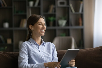 Joyful young woman resting on cozy couch with computer tablet in hands, watching funny photo or video content, web surfing information, spending free leisure time online in modern living room.