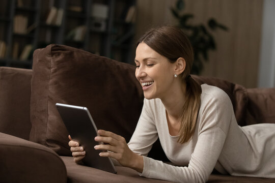 Cheerful young woman lying on sofa, using digital computer tablet, laughing watching funny photos or videos, communicating distantly in social network, playing games or shopping in internet store.