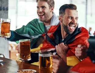 Happy young men covered in international flags toasting with beer while watching sport game in the pub