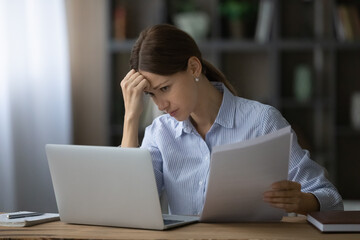 Stressed young businesswoman looking at computer screen, holding paper documents in hands, feeling...