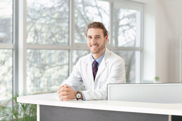 Fototapeta premium Smiling male doctor or medical professional in white lab coat at reception desk, healthcare and patient care