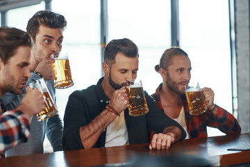 Group of handsome young men in casual clothing enjoying beer while sitting at the bar counter in pub