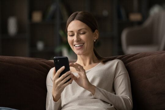 Sincere joyful young woman looking at cellphone screen, reading message with amazing news, having fun watching photo or video content online in social network, shopping or playing mobile game.