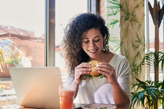 Smiling ethnic freelancer with delicious bagel sandwich against laptop indoors