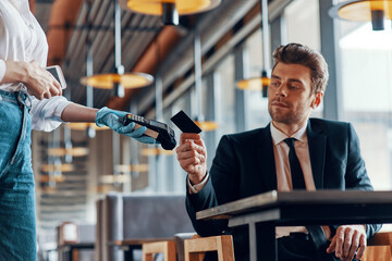Handsome young smiling man in full suit making a contactless payment while sitting in the restaurant © gstockstudio