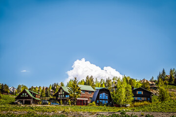 Group of log cabins and houses on mountain hillside with snow topped peaks in distance and pine and...