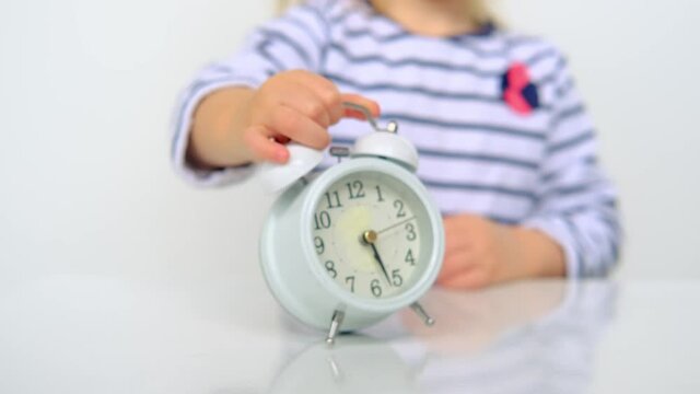 close-up the hand of a small child, a 2-year-old girl with white hair holds white alarm clock, concept of childhood, parenthood, time to school, get up on time