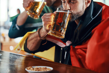 Young men covered in international flags enjoying beer while watching sport game in the pub