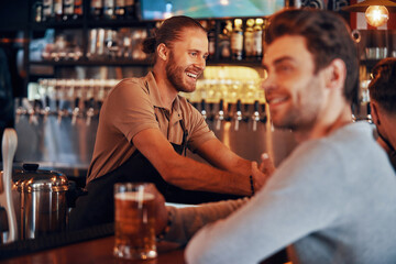 Cheerful bartender serving drinks while young smiling man enjoying beer on foreground in the pub
