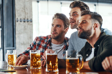 Three young men in casual clothing drinking beer and watching sport game while sitting in the pub