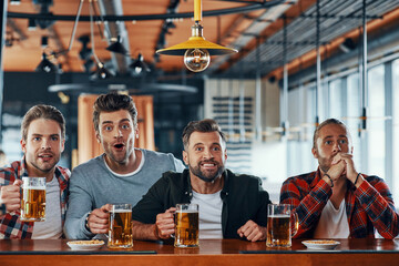 Cheering young men in casual clothing drinking beer and watching sport game while sitting in the pub