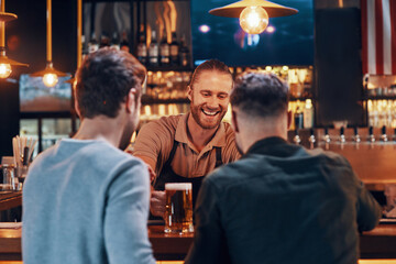 Cheerful bartender serving beer to young men while standing at the bar counter in pub