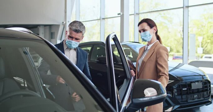 Confident caucasian female trader with tablet opening car door for the customer. Successful businessman choosing new automobile. Selling and business concept. Both in face masks.