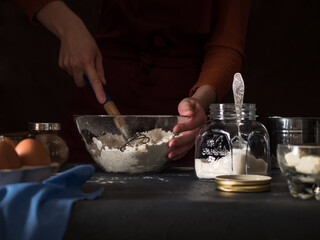 Cooking a cake. On the table are ingredients for making a pie (flour, eggs, cocoa, butter, sugar, salt), textiles. Kneading the dough with a whisk. In the background is a girl in an apron. 