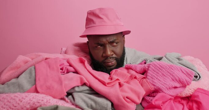 Dark skinned bearded man sticks head from pile of dirty clothes prepares for doing laundry wears panama poses against background has serious expression then smiles positively. Sorting out clothing