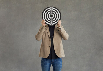 Unknown businessman hid his face behind a darts board while standing on a gray background. Concept...