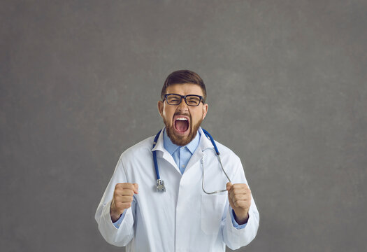 Studio portrait of young doctor stressed by problem at work going postal and shouting. Angry frustrated mad crazy furious general practitioner clenching fists and screaming isolated on gray background