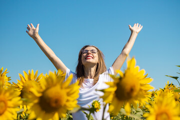 Young woman in a white t-shirt and glasses with raised up hands on a sunflower field on a summer sunny day