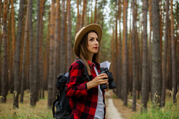 Young woman tourist in a hat, red plaid shirt holds a map and binoculars in the forest