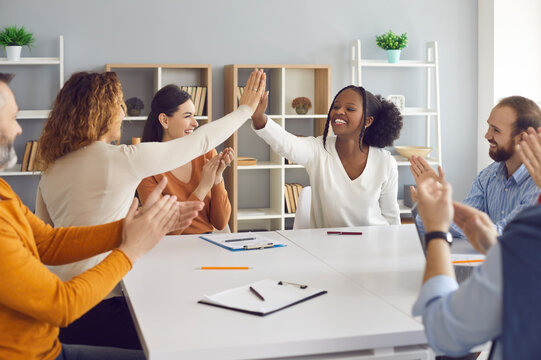 Teams Of Happy Diverse Business Professionals Making A Deal. Female Group Leaders Celebrating Successful Teamwork. Two Smiling Young Women High Five Each Other Sitting At Office Table In Work Meeting