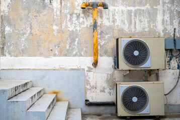Two old air conditioners are mounted on an old wall beside the stairs and are attached to an old...