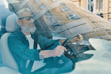 Active senior man in casual clothing enjoying car ride while sitting on front seat of the car