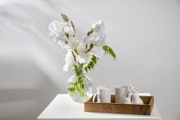 A bouquet of three white irises and a fern in a transparent vase on the table. Two ceramic tea in the wooden tray. Breakfast