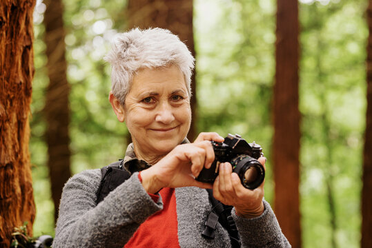 portrait of 70 year old senior woman looking at camera and holding with her hands a photo camera in the forest. retired adventurer