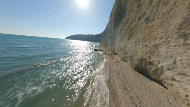 Rapid fast movement fpv sport drone over natural beach sea coast near high mountain rock at summer sunny day. Aerial shot view flying at seashore with calmness waving water and clear blue sunny sky