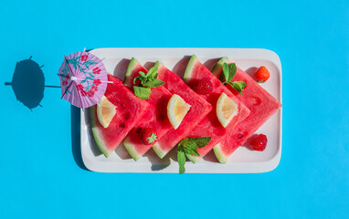 Watermelon slices with berries and lemon. 
Watermelon slices with berries and lemon in a plate lie in the middle on a blue background, close-up top view.