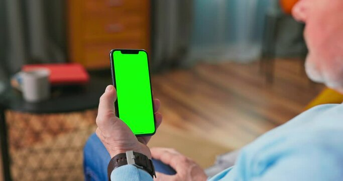 Adult Old Man at Home Uses Green Mock-up Screen Smart Phone. He is Sitting On a Couch in His Cozy Living Room. Man clicks one time on the screen cell phone and browses the content.