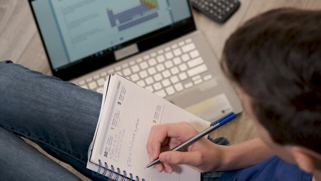 Caucasian teenager boy in casual wear using laptop and copybook for distance education or online learning in home. Boy sitting on floor, writing concept, modern e-learning. Educational platform.