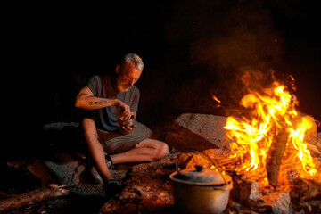 Mature man, tourist eating dinner while sitting alone near campfire in the wild