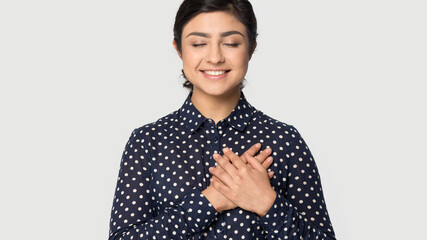 Happy grateful young Indian woman isolated on grey studio background hold hands at heart chest show...