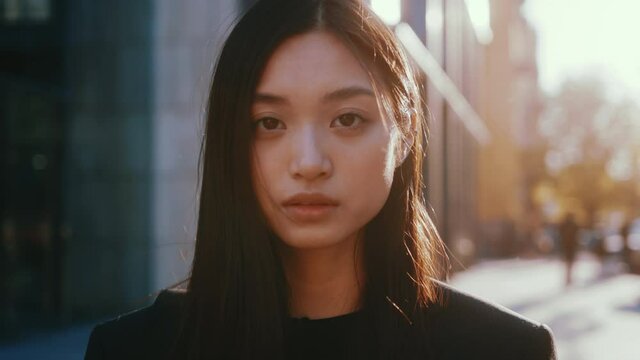 Portrait of nice mixed-race asian young beautiful woman with straight black hair posing to camera outside. Urban people, cityscape, sunlight. Females.