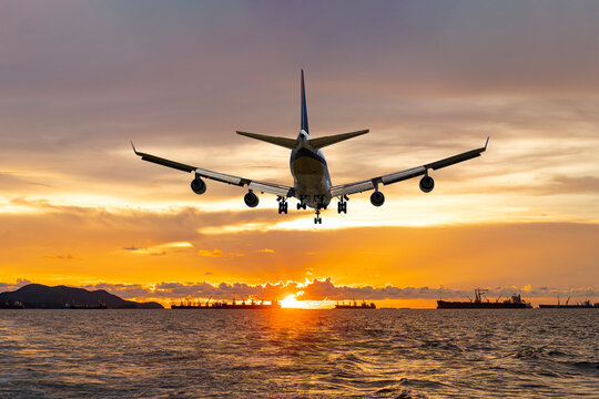 rear image commercial passenger aircraft or cargo airplane fly over coast of sea after takeoff from airport in evening with golden sunset seascape view and bulk cargo ships anchored at horizon line