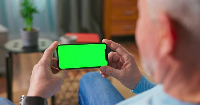 Older man at Home Resting on a Couch Using with Green Mock-up Screen Smart Phone in Horizontal Landscape Portrait Mode. Woman Using Tablet Device, Browsing Internet, Watching Content, Videos.
